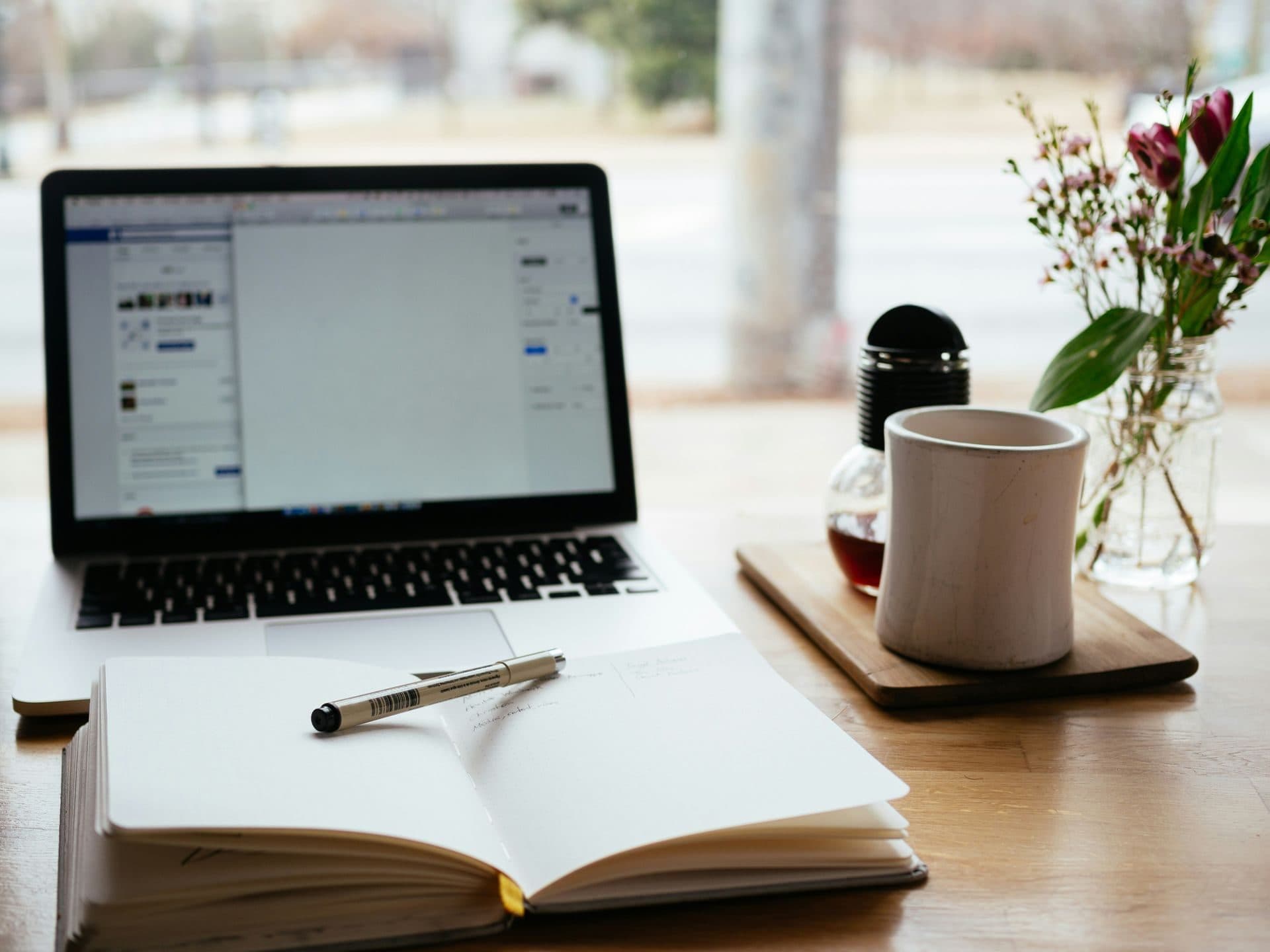 A desk with a laptop, notebook, coffee and flowers