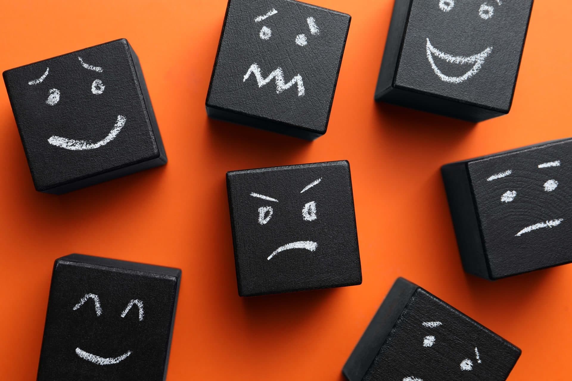 Black cubes with different hand-drawn facial expressions on an orange background, representing shifting moods