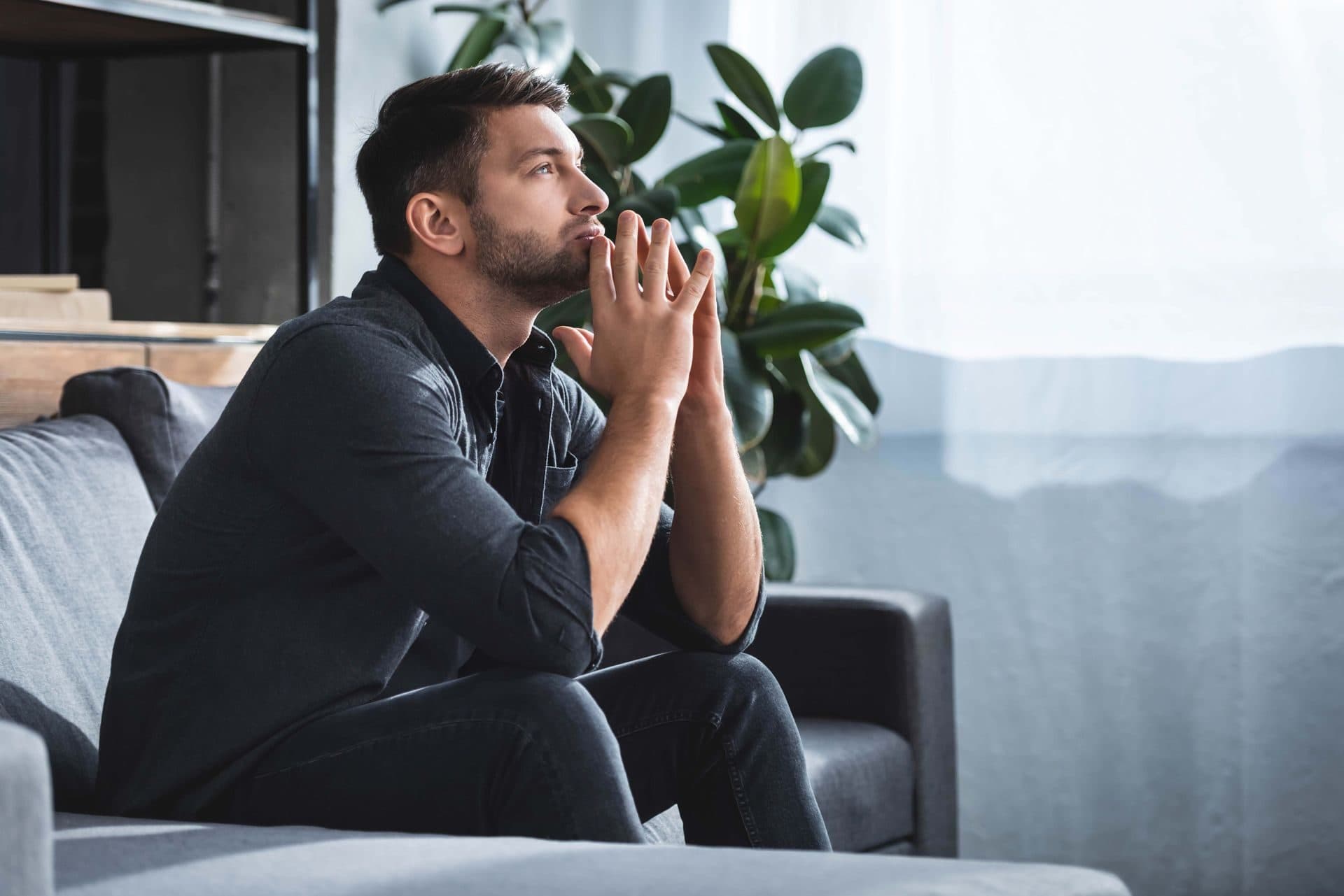A man sitting thoughtfully on a couch with his hands clasped, reflecting during a therapy session