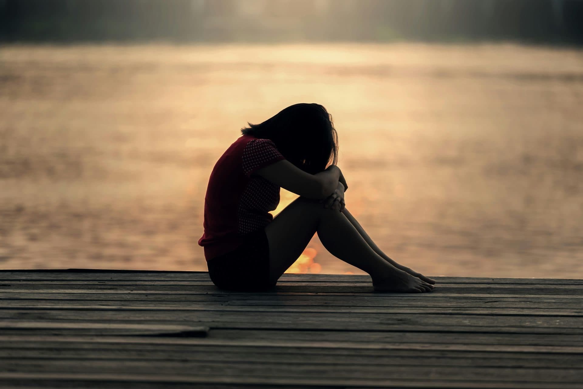 A woman sitting alone on a wooden dock at sunset with her head resting on her knees, representing grief
