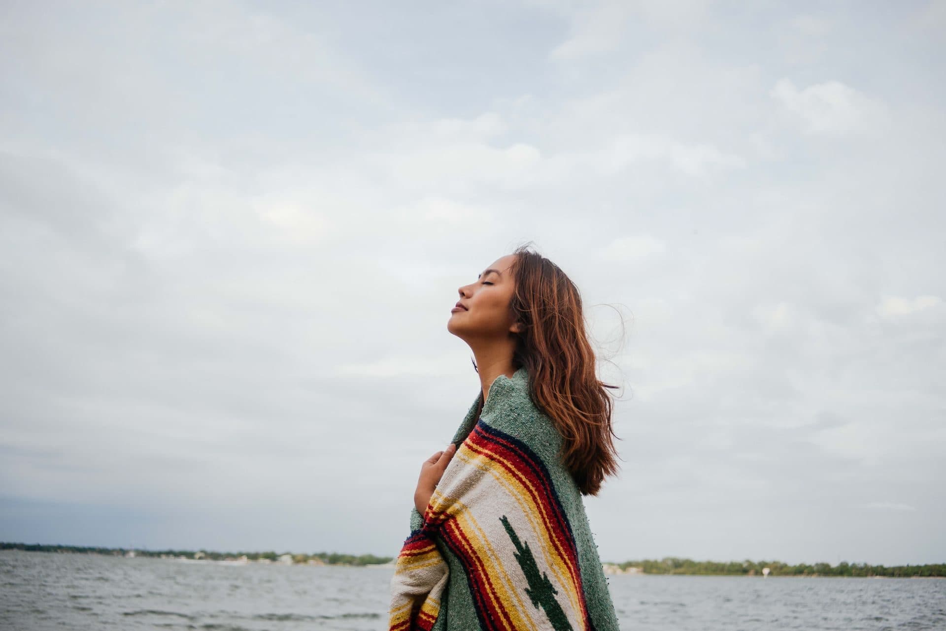 A woman wrapped in a blanket looking peacefully at the sky beside calm water